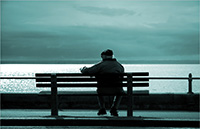 Elderly man sitting on a bench looking out over a lake.